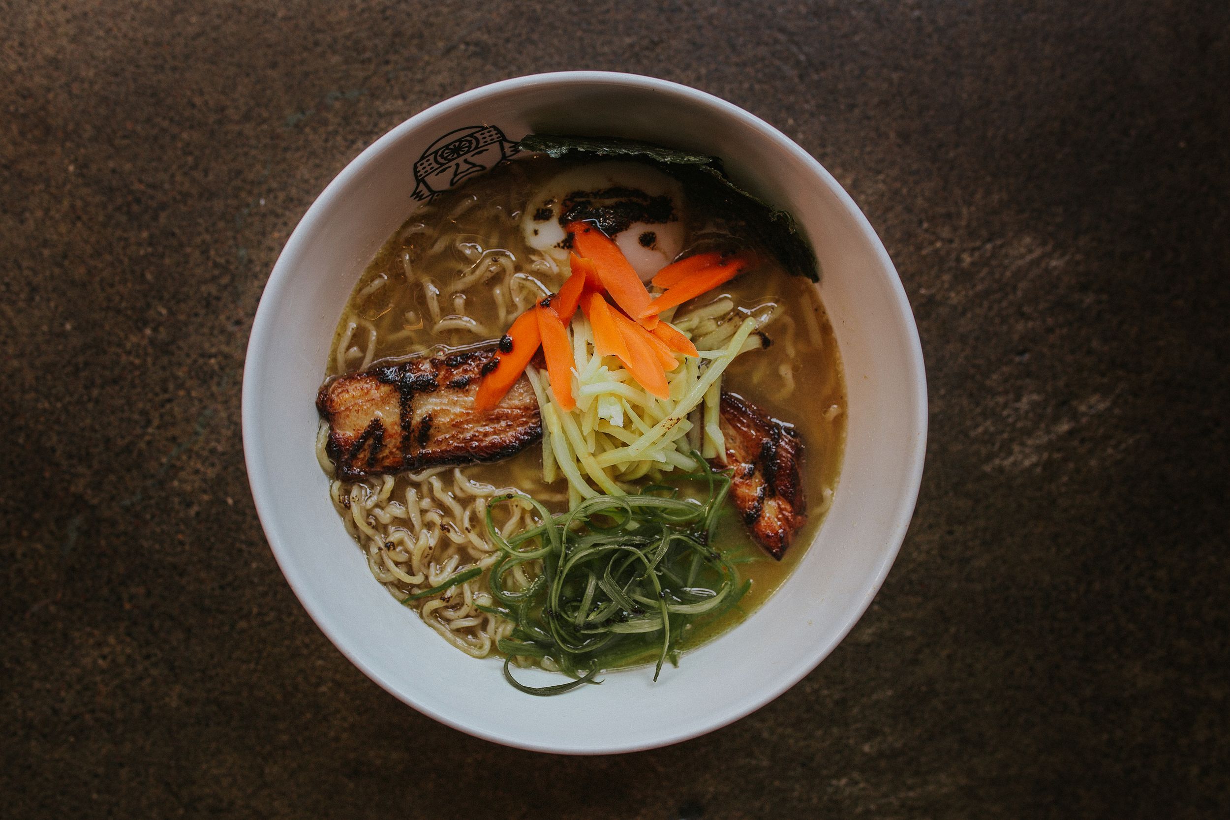 A steaming bowl of ramen noodle soup with colorful vegetables, garnished with sliced carrots and scallions in a white bowl.