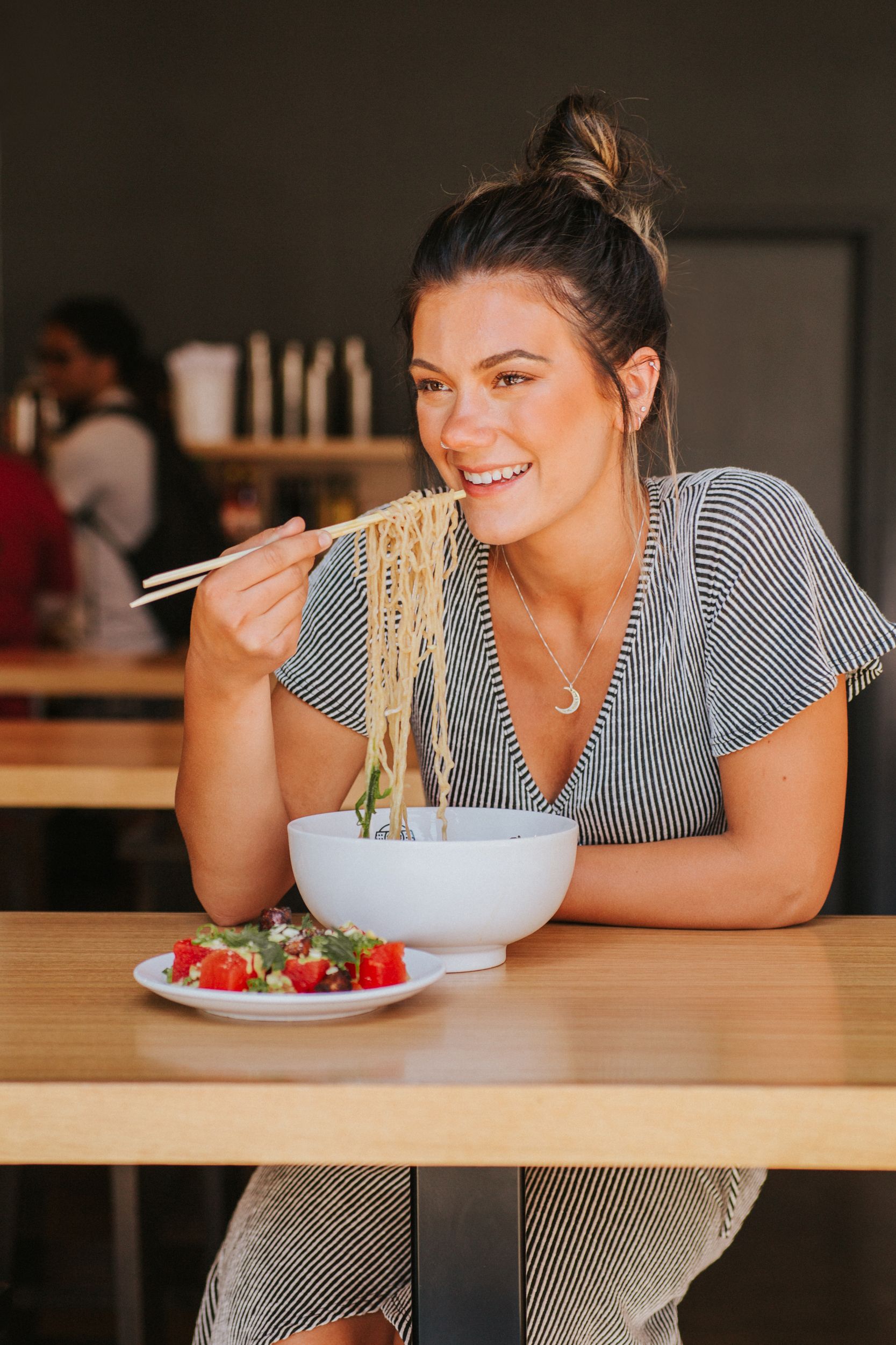 Smiling woman in striped shirt enjoys eating ramen noodles from a bowl while sitting at restaurant table with strawberries.