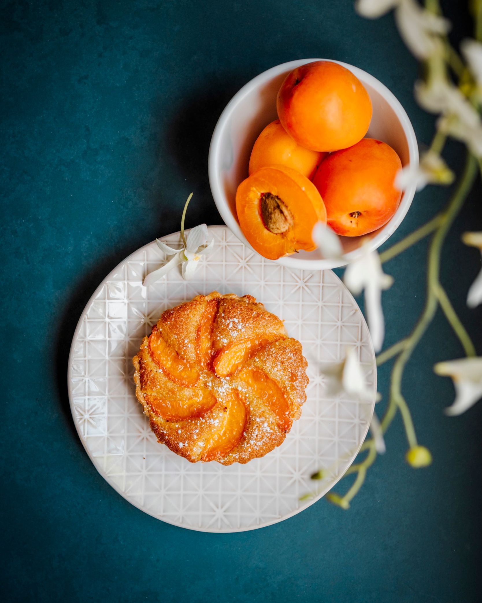 Overhead view of a flaky pastry with spiral design on white plate beside bowl of fresh oranges on teal surface.
