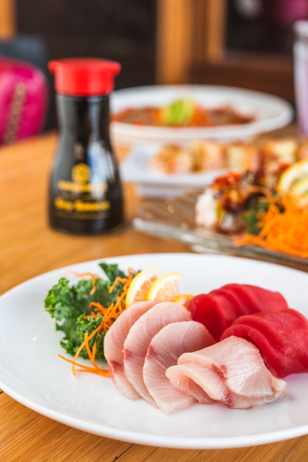 Fresh sashimi slices of tuna and fish served on white plate with garnish and soy sauce bottle in background.