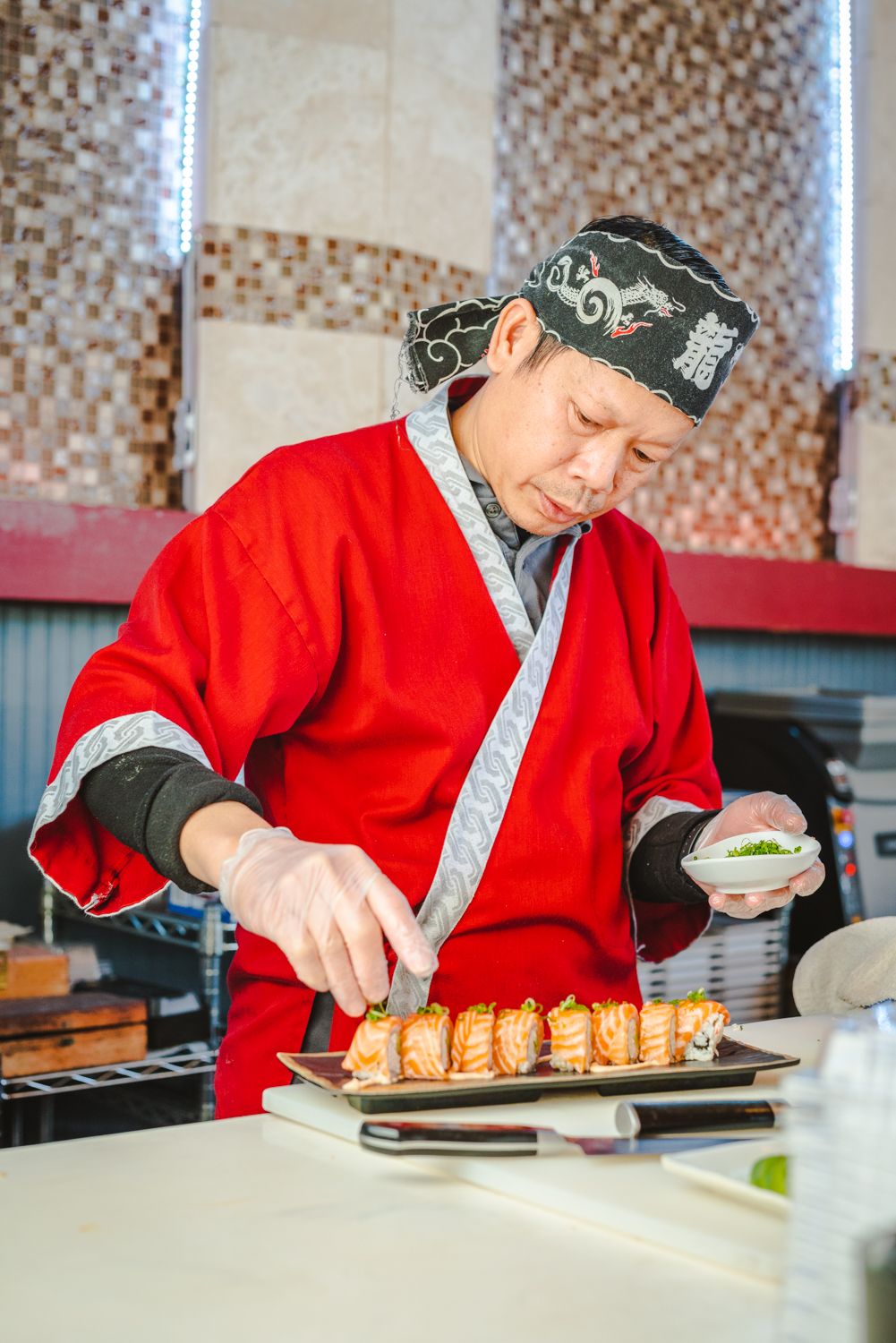 A chef in red traditional Asian attire carefully prepares sushi rolls at a restaurant counter.