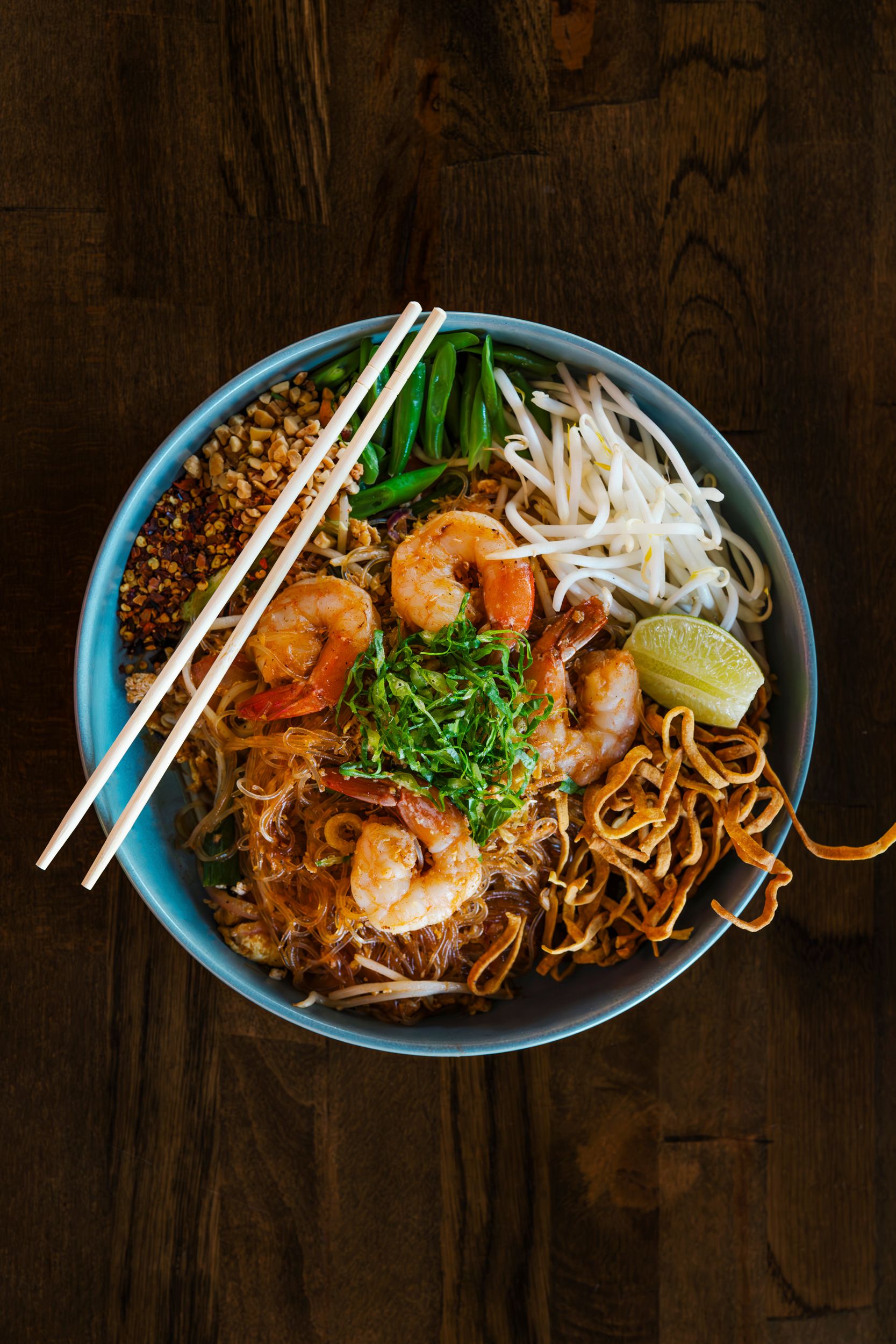 Overhead shot of shrimp pad thai noodles in blue bowl with bean sprouts, lime and chopsticks on dark wooden table.