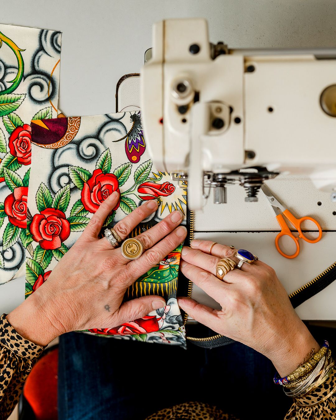Hands adorned with gold rings work on a colorful floral fabric next to a vintage white sewing machine and orange scissors.