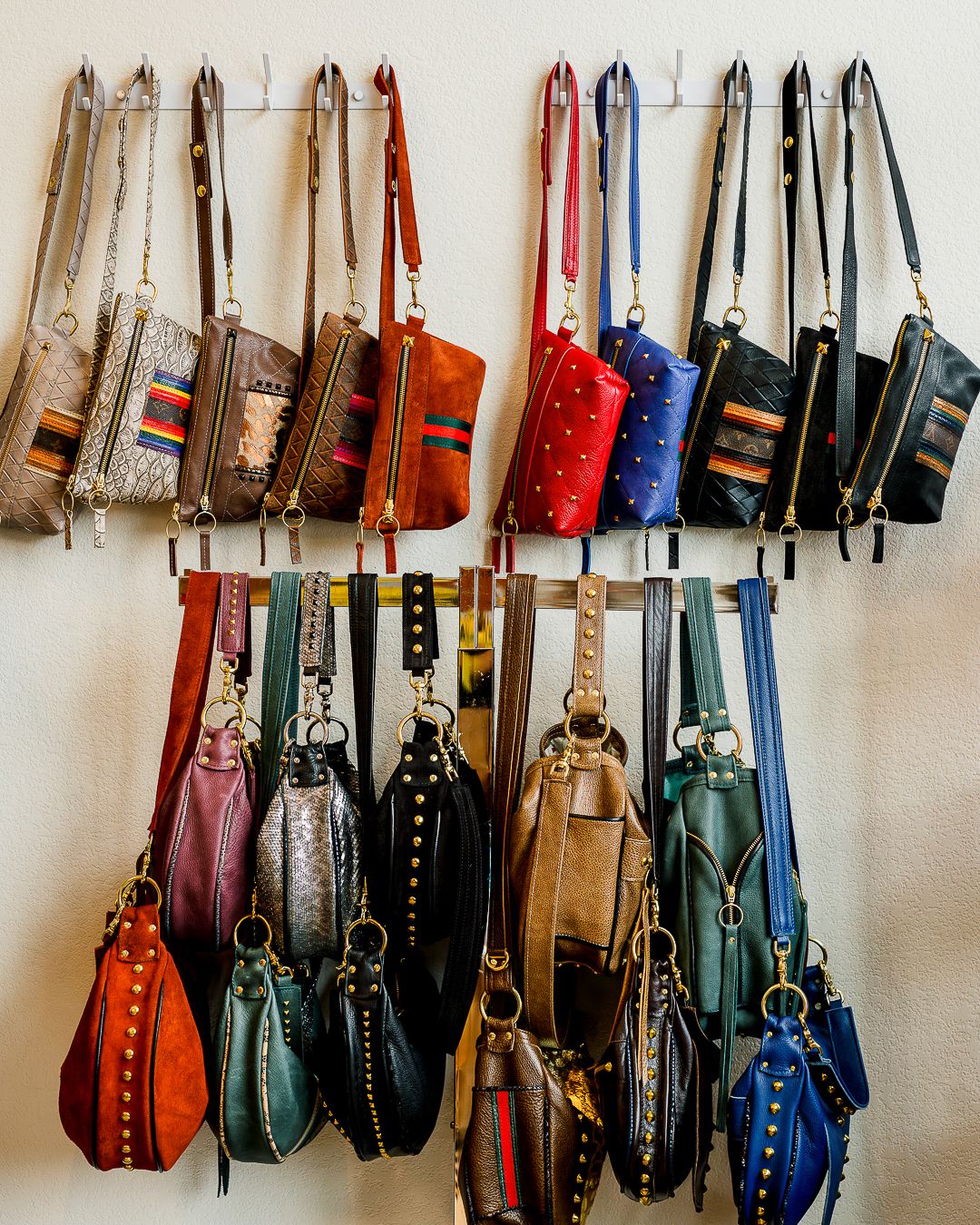 Collection of colorful handbags and purses hanging on white wall display with leather and fabric styles in various sizes.