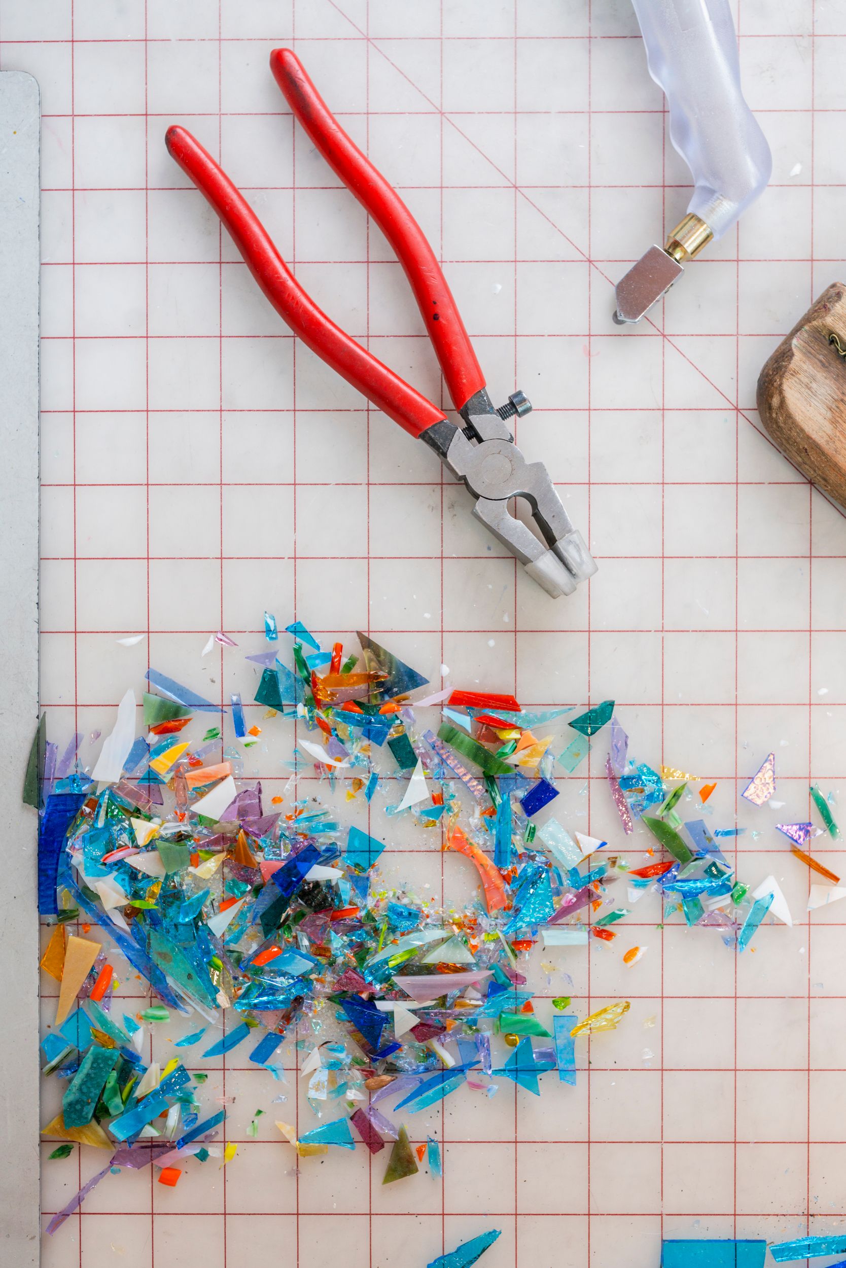 Red handled pliers with scattered colorful mosaic glass pieces on a cutting mat background.