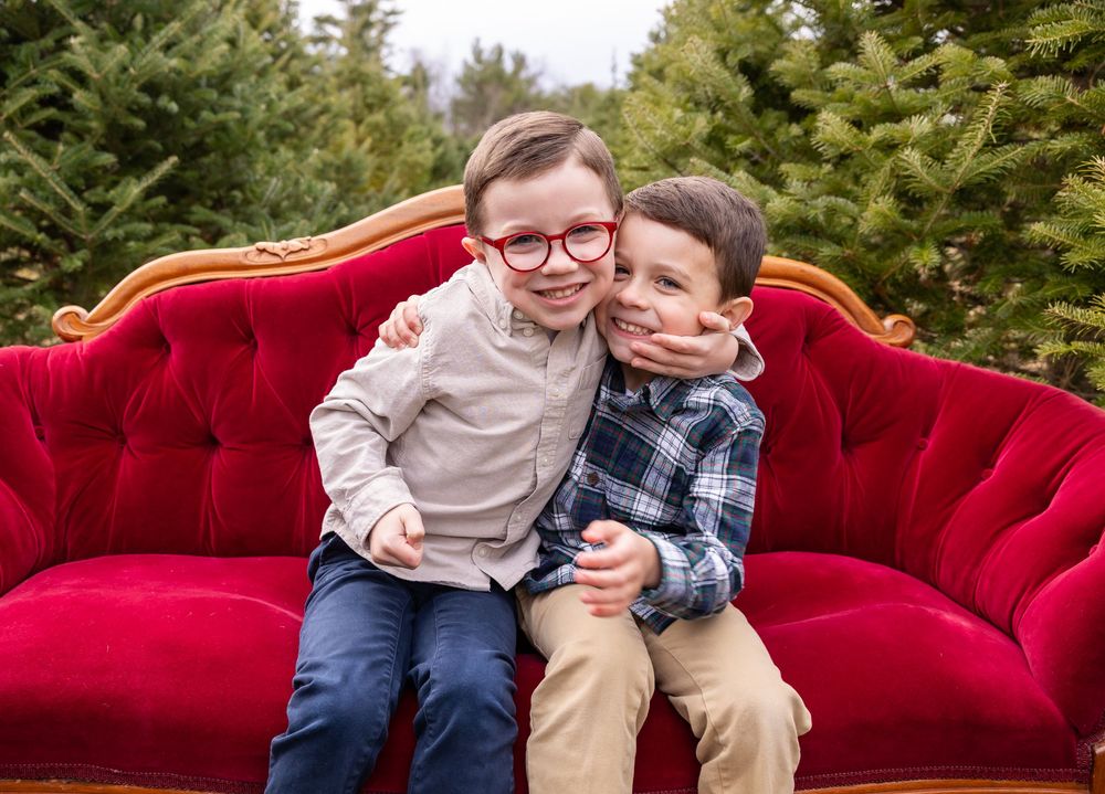 Two children share a joyful moment while sitting on a plush red velvet couch surrounded by Christmas trees.
