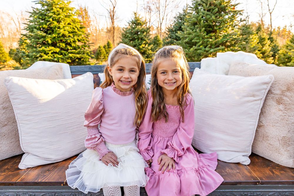 Two young girls in pink dresses sitting on an outdoor couch with white pillows.