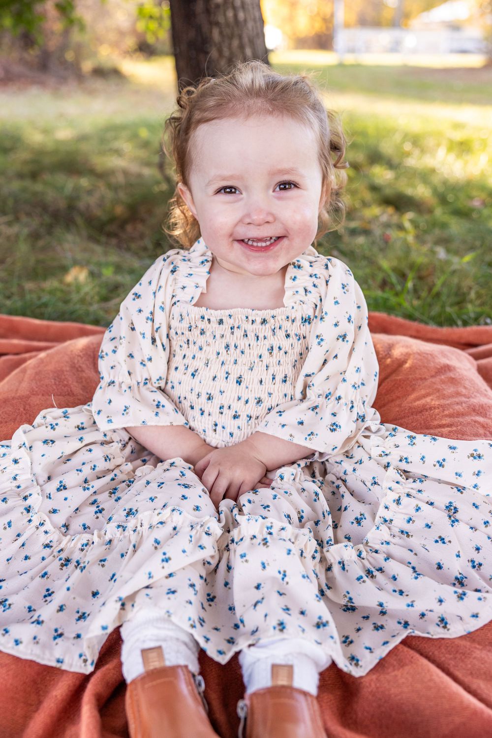 Young child in floral dress sitting on orange blanket outdoors during autumn.