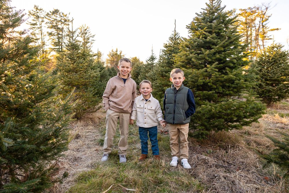 A group of people stand together among evergreen trees at a Christmas tree farm during golden hour.