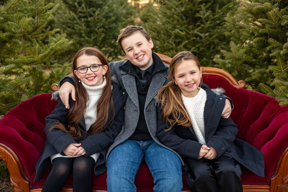 A family poses together on a burgundy couch against a backdrop of evergreen trees during a holiday photo session.
