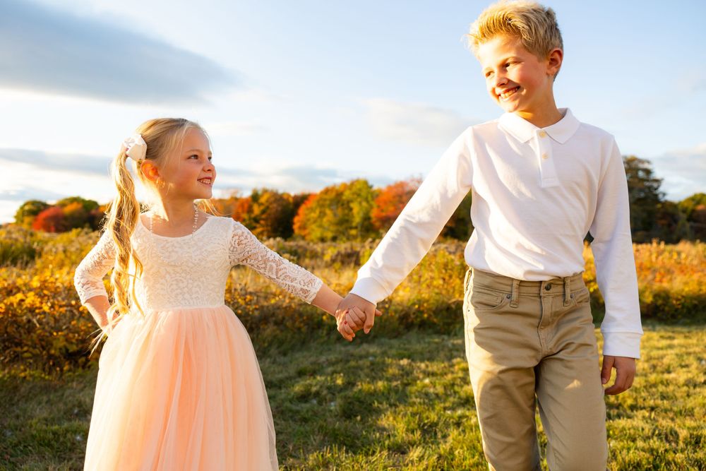 Two children holding hands in a field during golden hour with autumn foliage in the background.