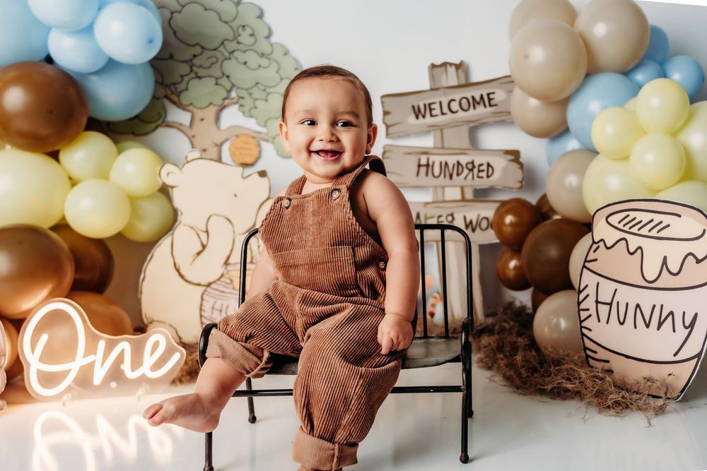 First birthday celebration setup with brown overalls, Winnie-the-Pooh theme decor, pastel balloons and welcome sign.