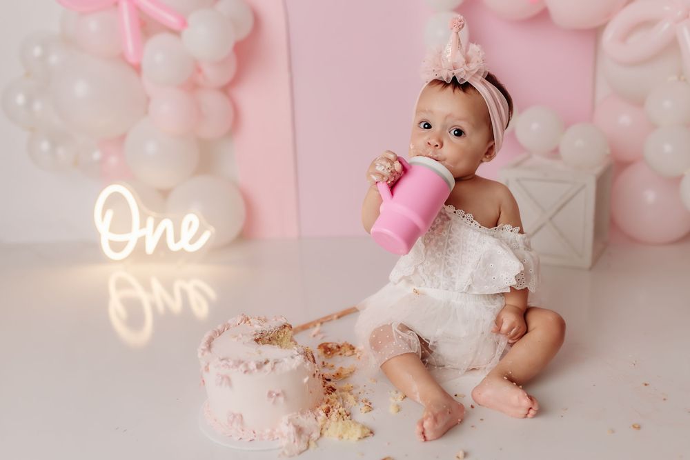 A pink and white baby themed cake smash photography session with floral decorations and balloons in the background.
