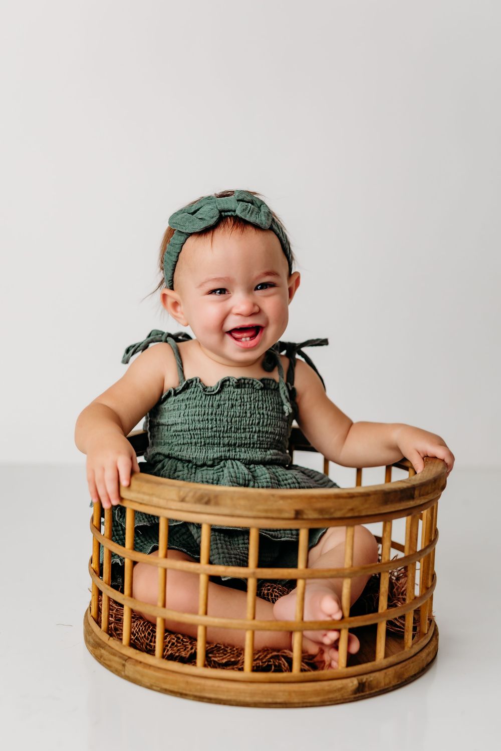A small child wearing green clothing sits and laughs joyfully while playing in a round wooden basket container.