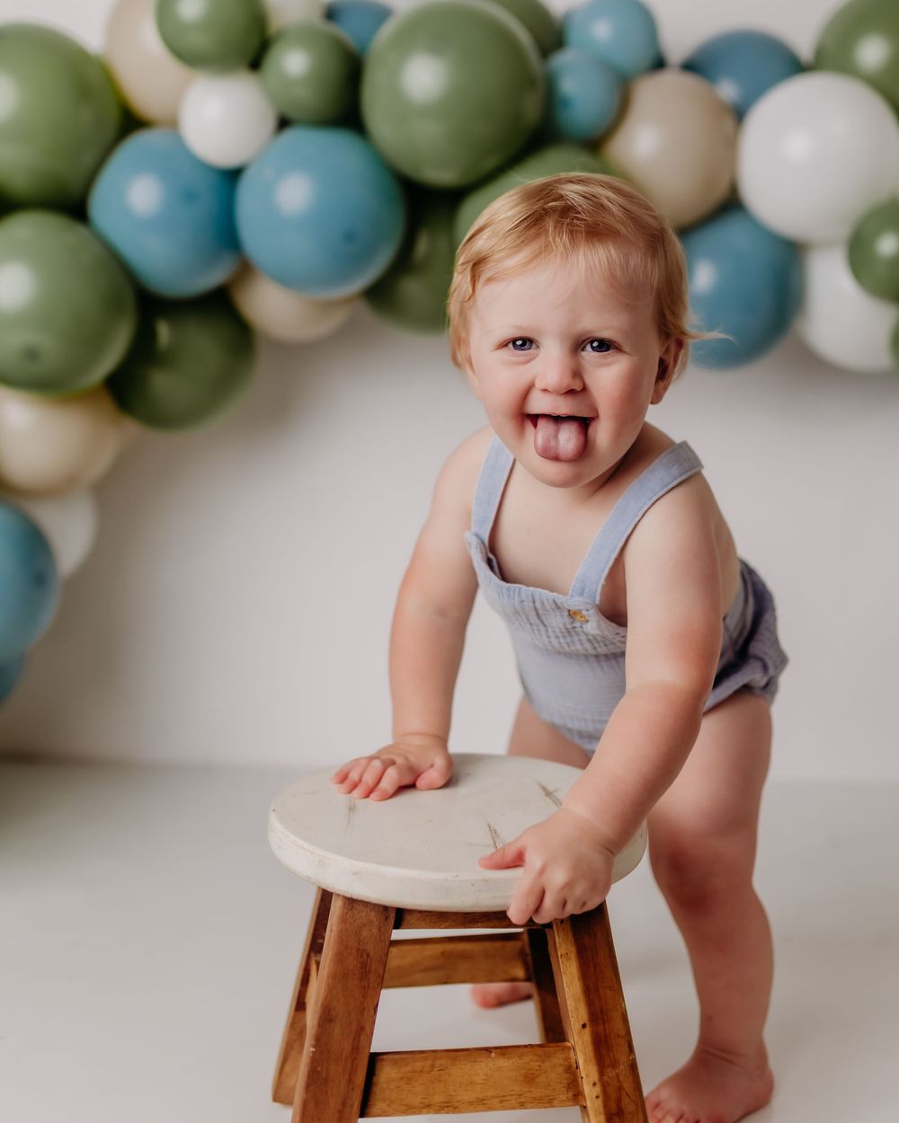 Blue, white and olive green balloon garland decorates backdrop behind wooden stool for first birthday photo session.