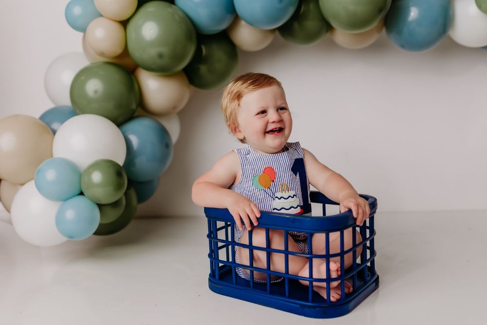 A small child sits inside a navy blue plastic crate against a wall decorated with sage green and blue balloon garland.