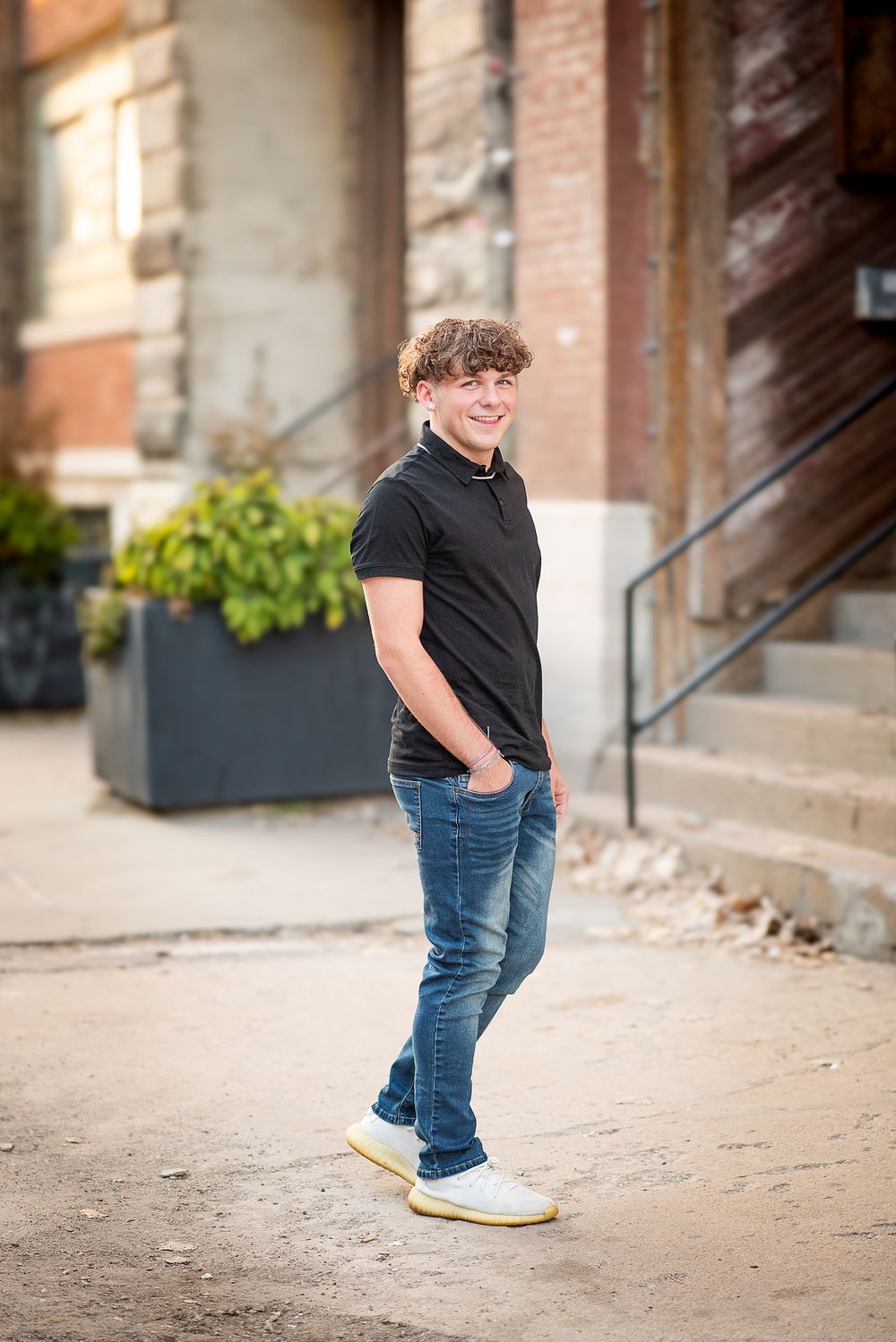 Smiling teenage boy with curly hair wearing black polo and jeans posing outdoors near brick building steps.
