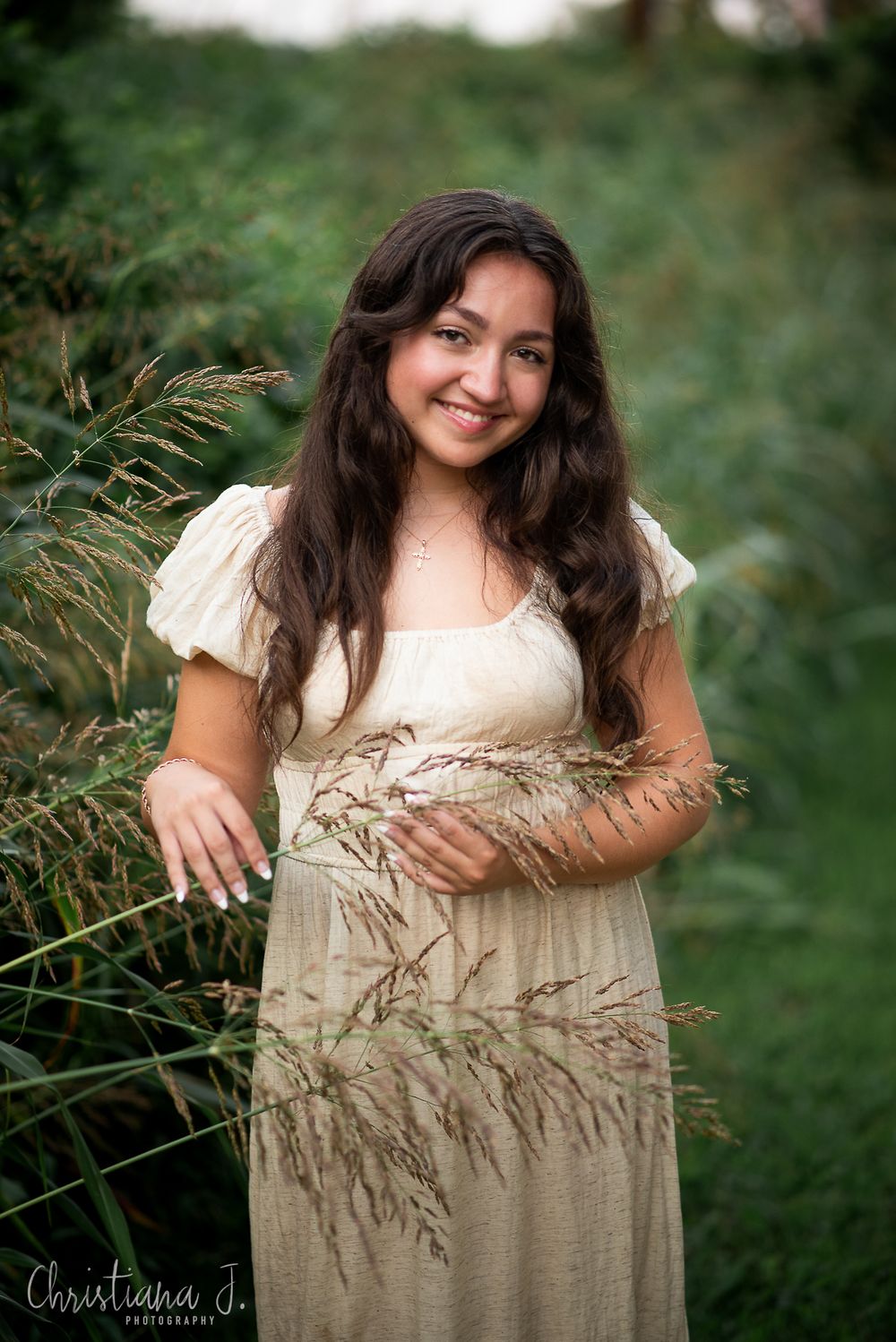 smiling brunette high school senior girl against greenery and tall grasses wearing a natural boho dress in olden hour light in kansas city