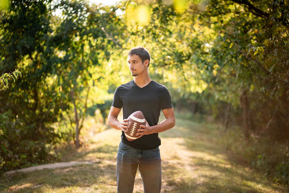 Young man in black t-shirt holding a football on a sunlit tree-lined path outdoors.