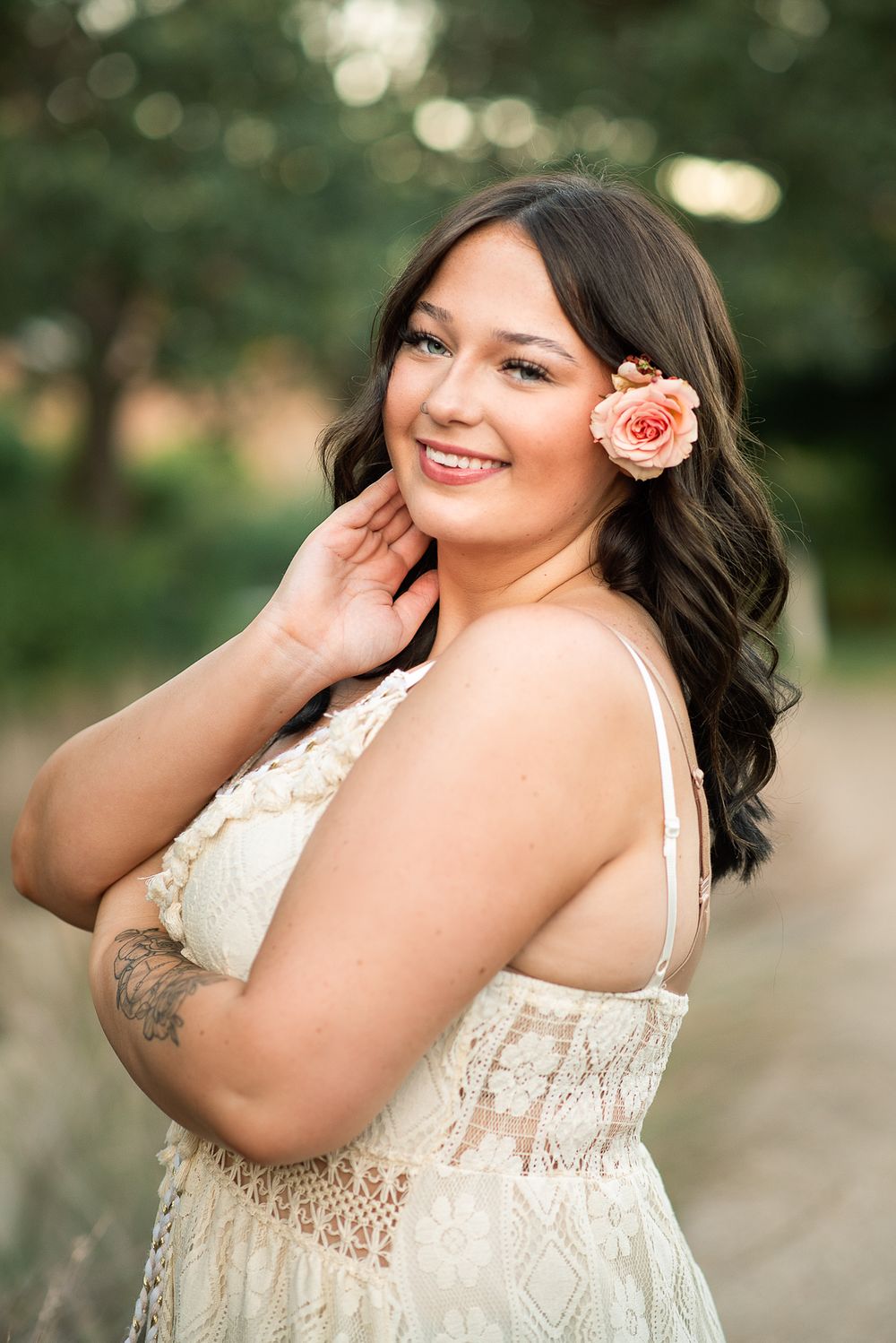 Smiling woman in white lace dress with peach flower in dark wavy hair, posing outdoors in soft natural light.