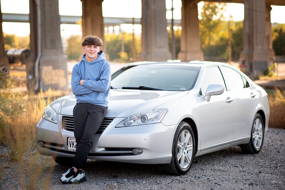 Teen boy in blue hoodie poses leaning against a silver Lexus sedan outdoors near a bridge.