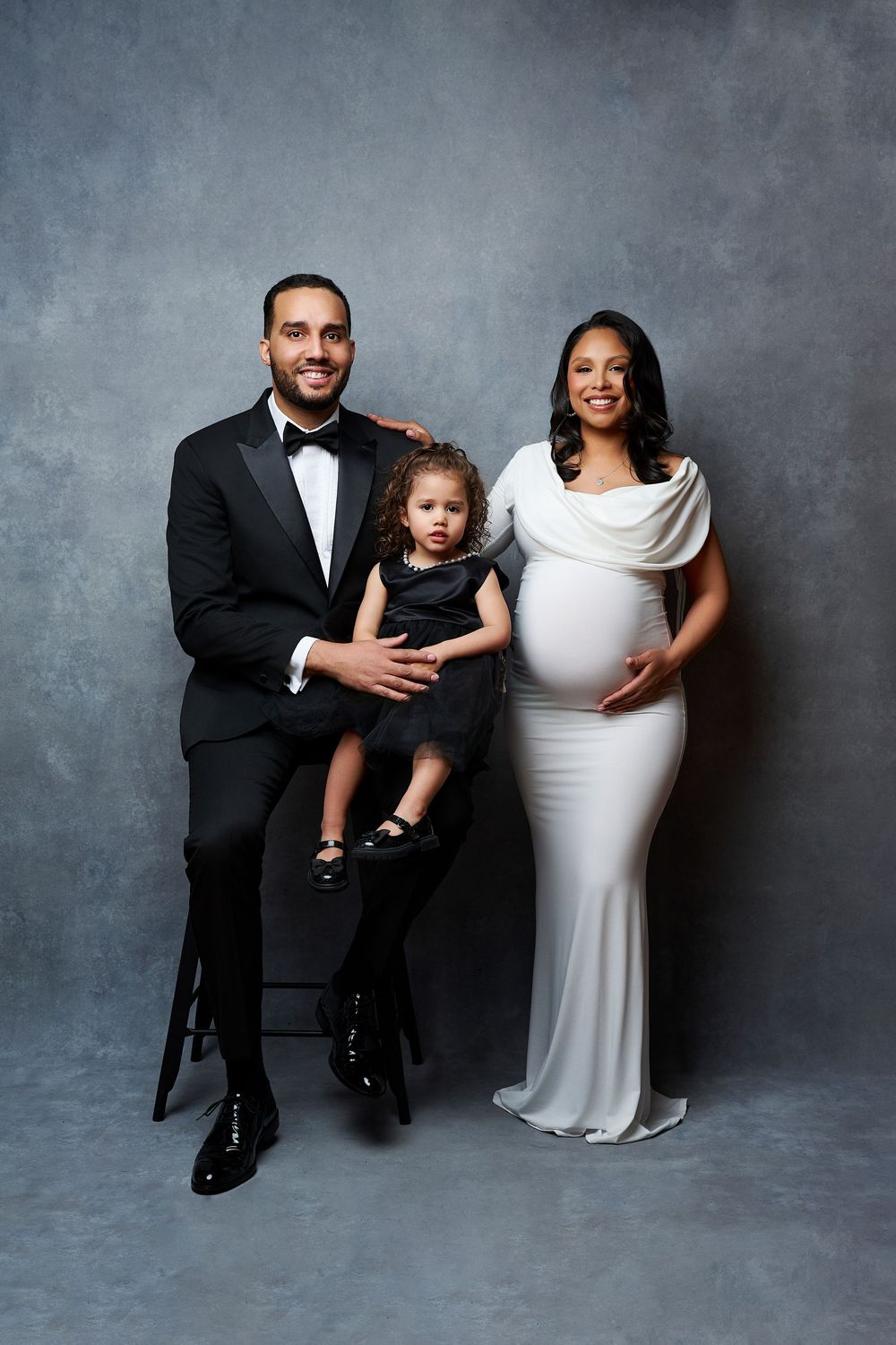 Elegant family maternity photo with man in tuxedo, toddler in black dress, and pregnant woman in white gown.