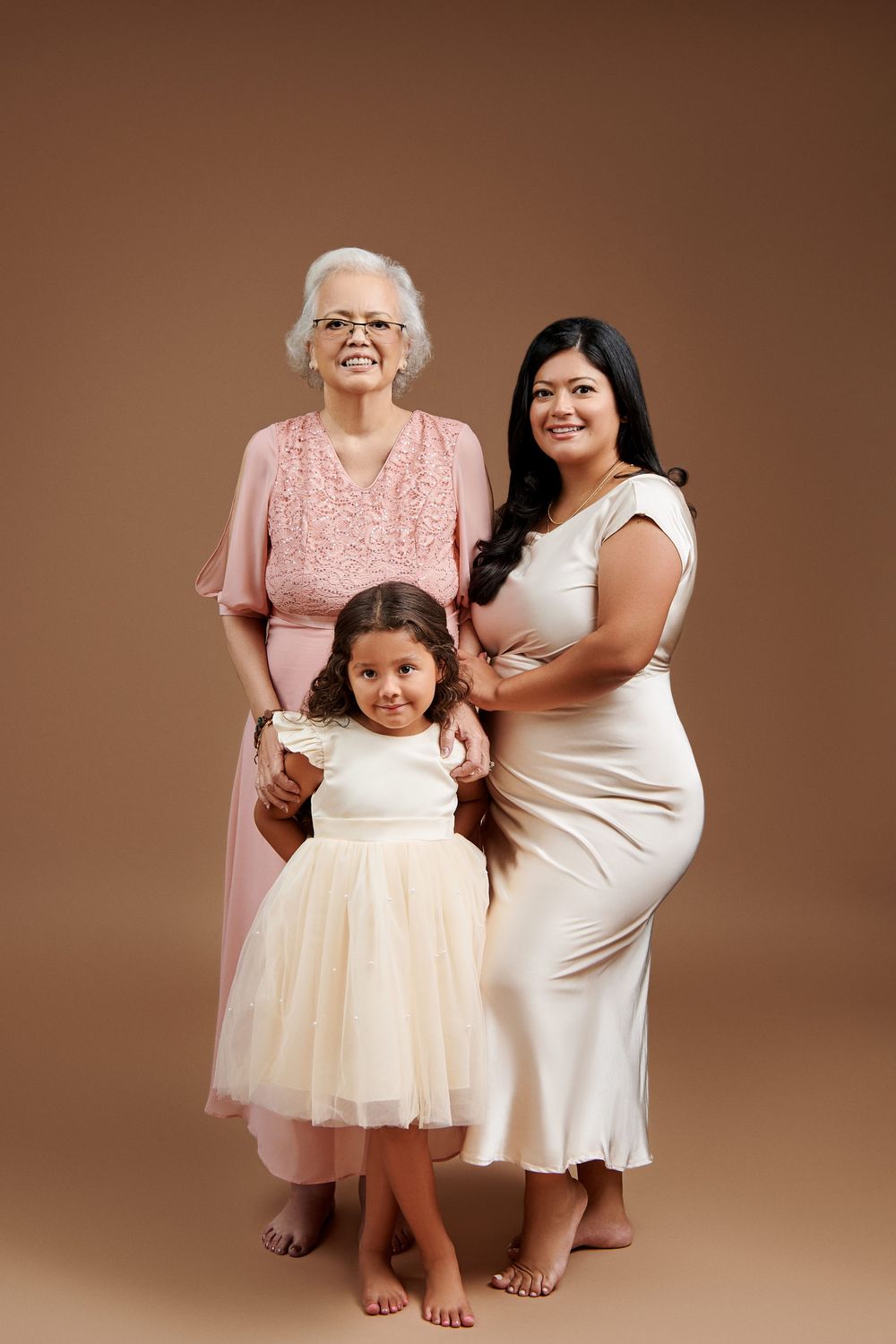 Three generations pose together in matching pink and cream colored dresses against a brown studio backdrop.
