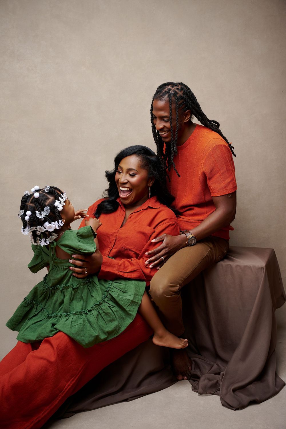 A joyful family portrait with a laughing mother, father with braids, and young daughter in green dress.