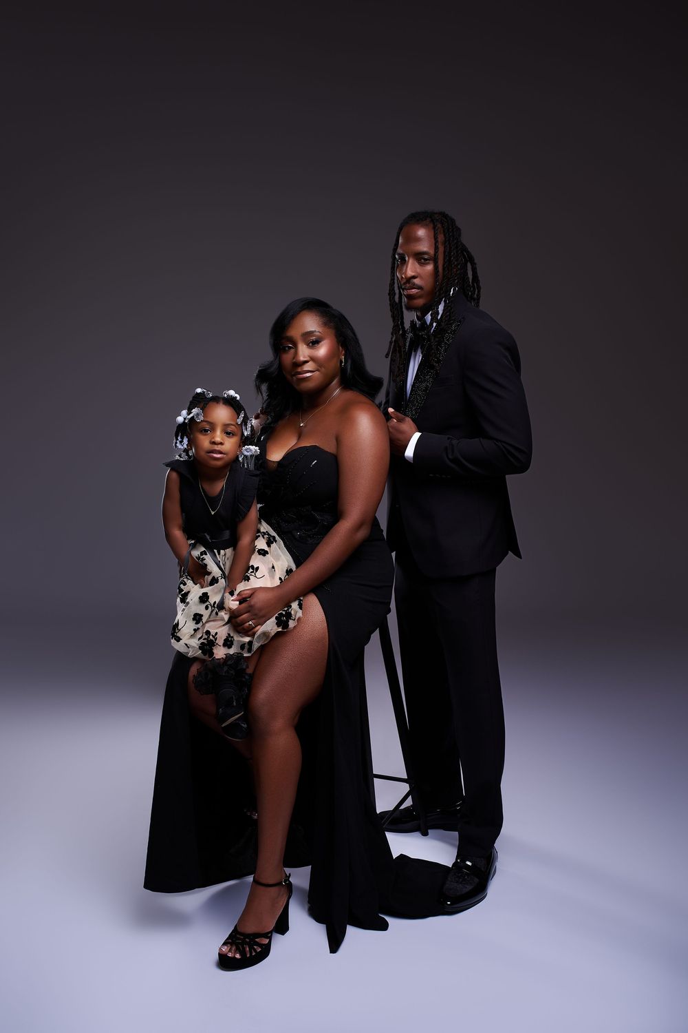 A family of three poses elegantly in a studio, wearing formal black attire against a dark gray background.