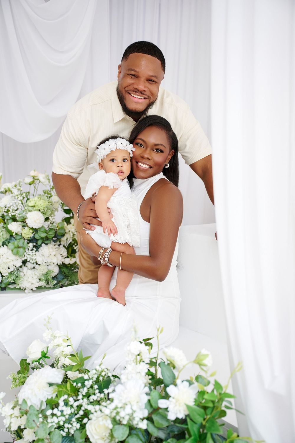 Happy family portrait with parents in white attire holding their baby girl, surrounded by white floral arrangements.