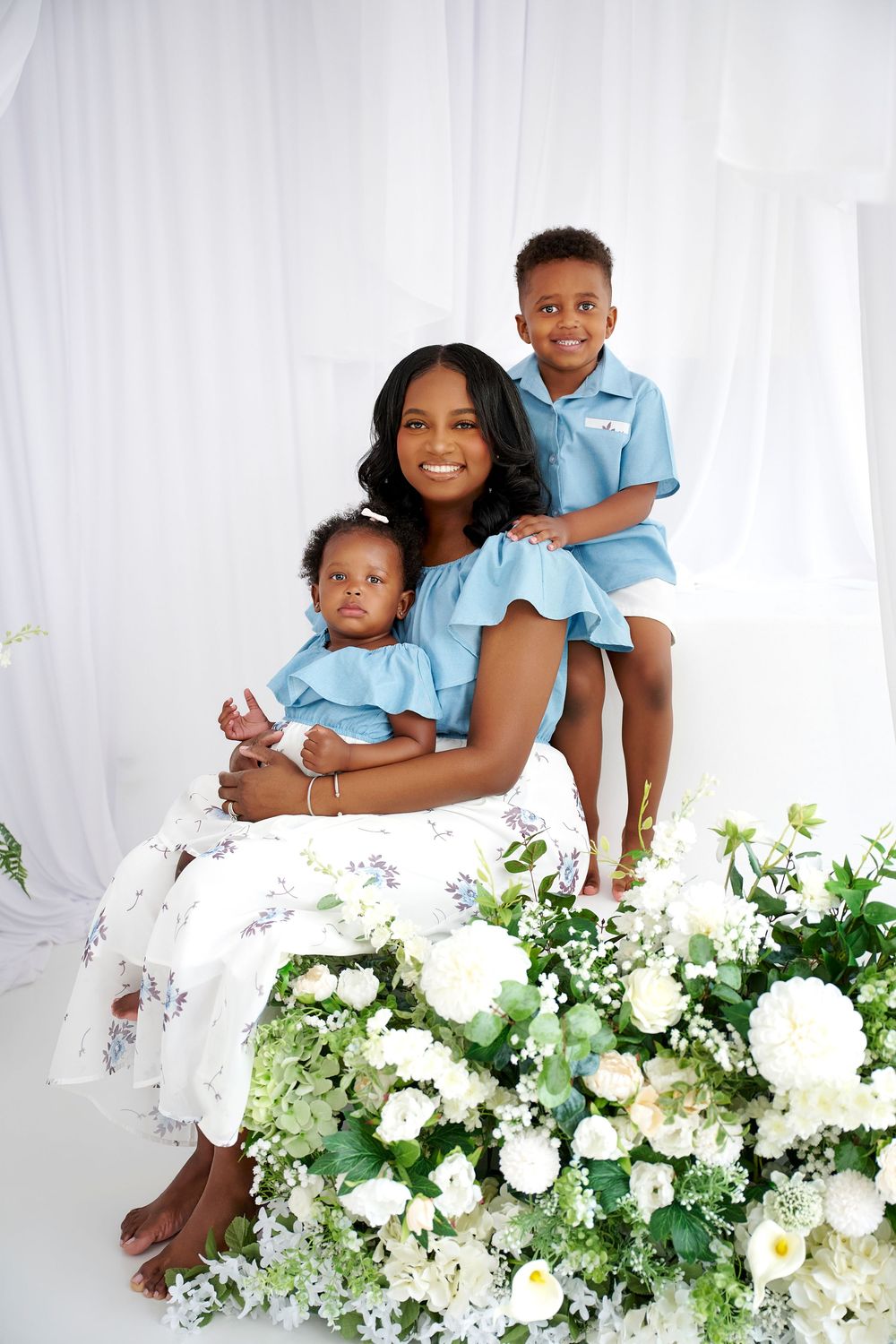A smiling mother in blue sits with her two young children amid white flowers and greenery in a bright studio portrait.