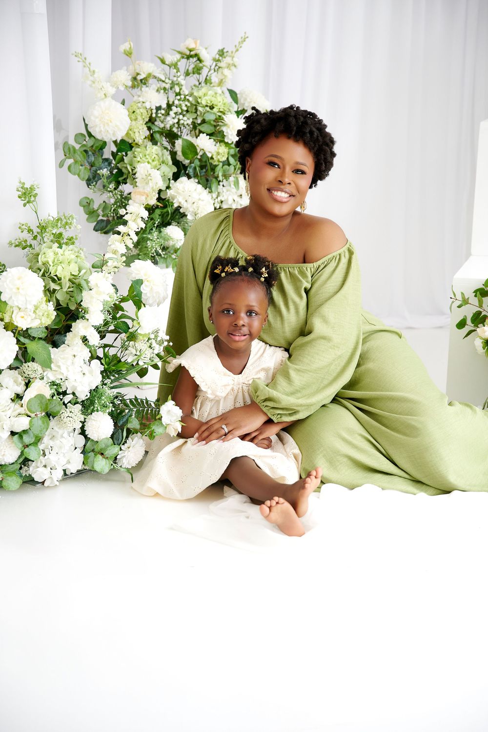 A Black mother in a green dress smiles with her young daughter in white, surrounded by white floral arrangements.