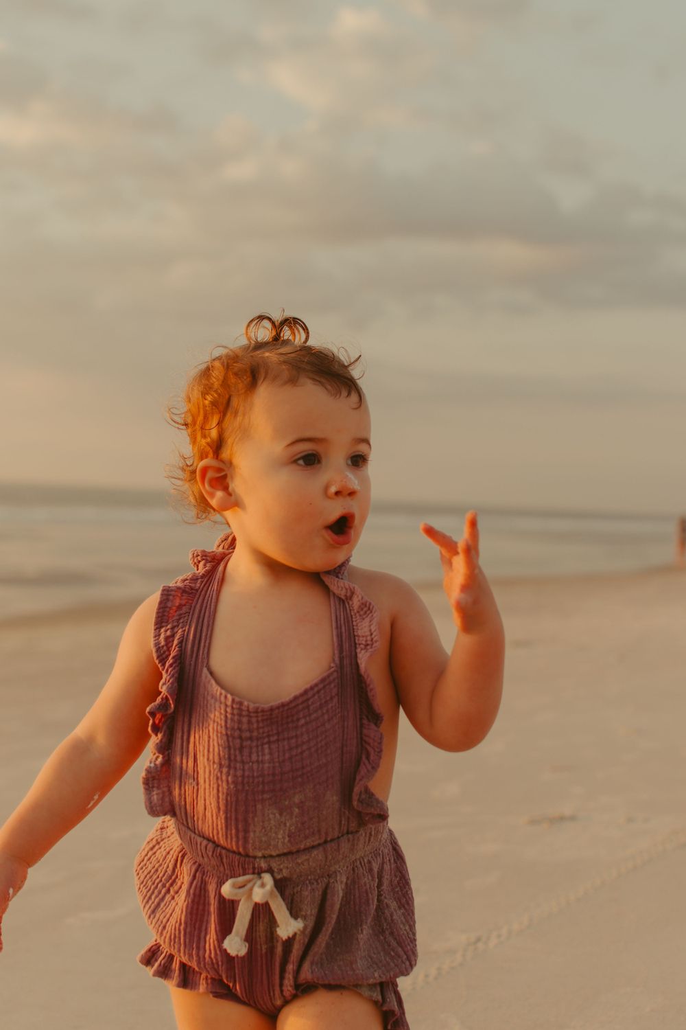 Toddler in dusty rose romper playing on beach at sunset.