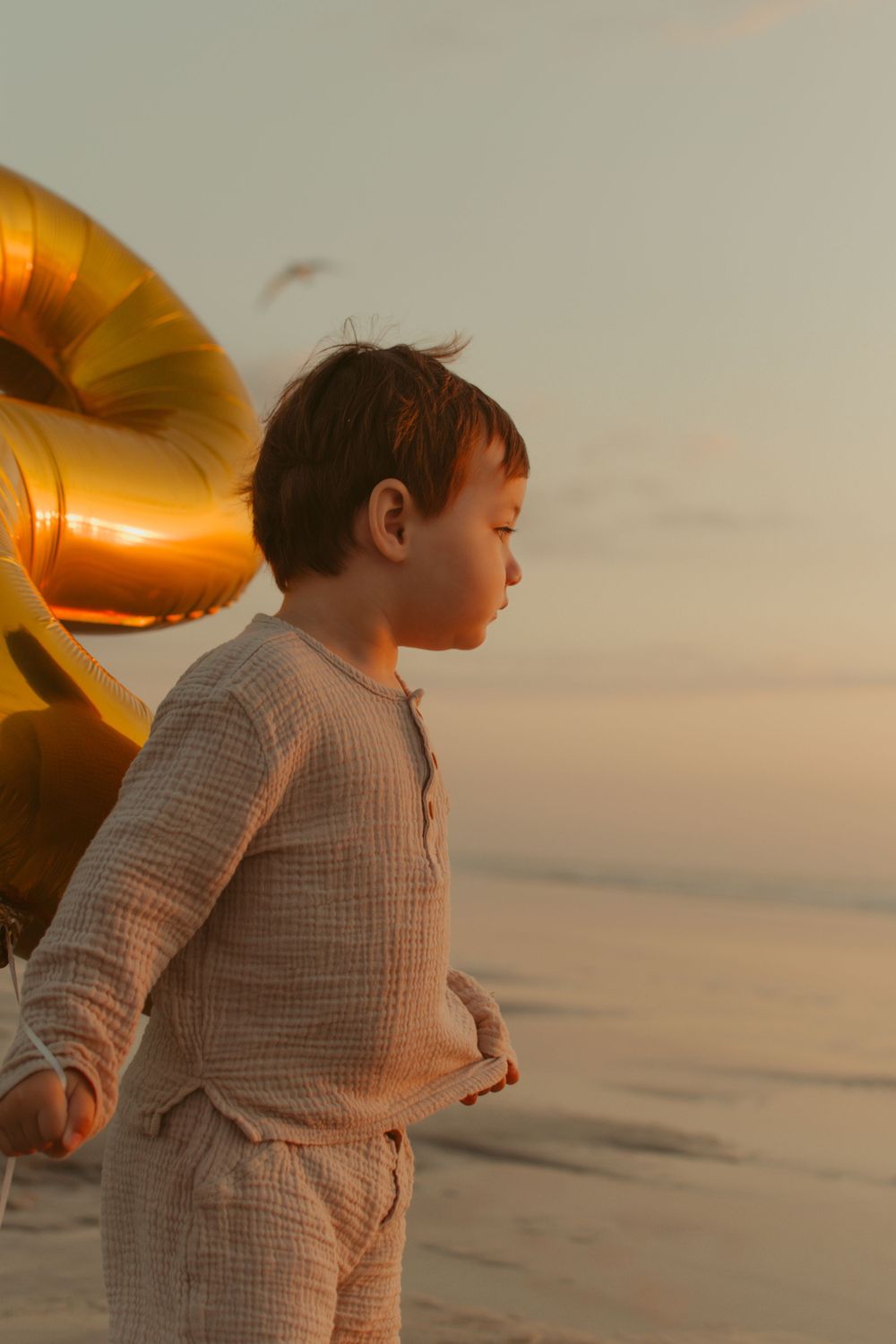 Child in beige outfit standing on beach with golden balloon in background at sunset.