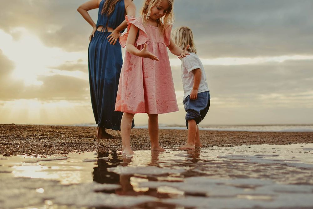 Three people standing in shallow ocean water at sunset wearing colorful dresses.