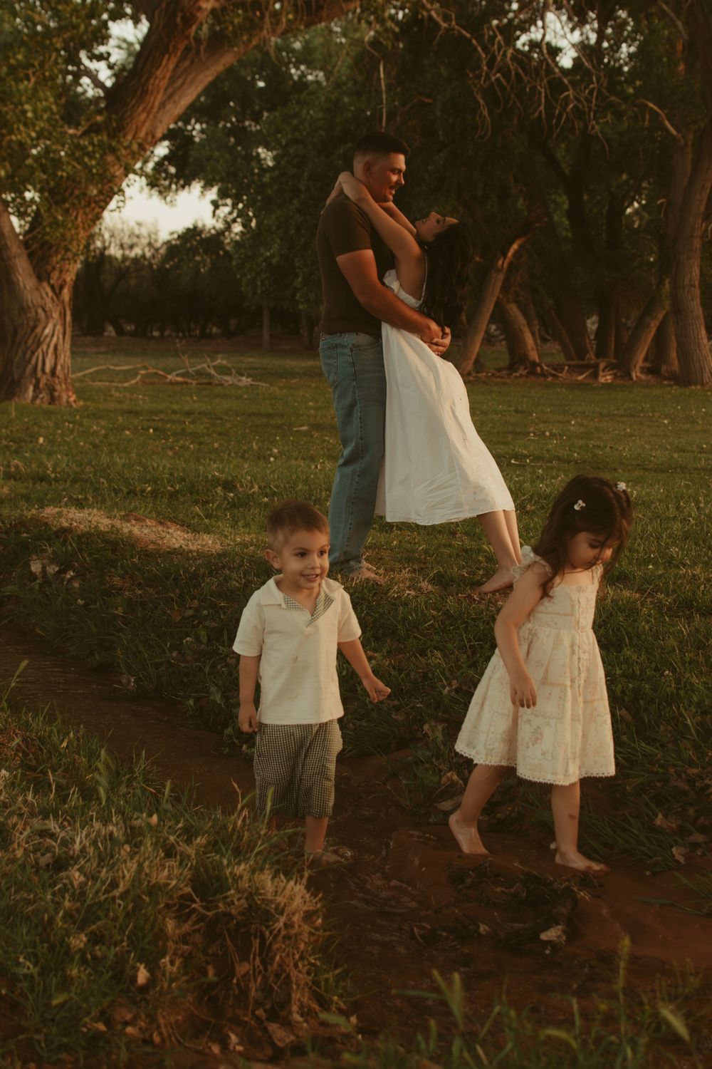 Family enjoying sunset together under trees in a park with children playing.