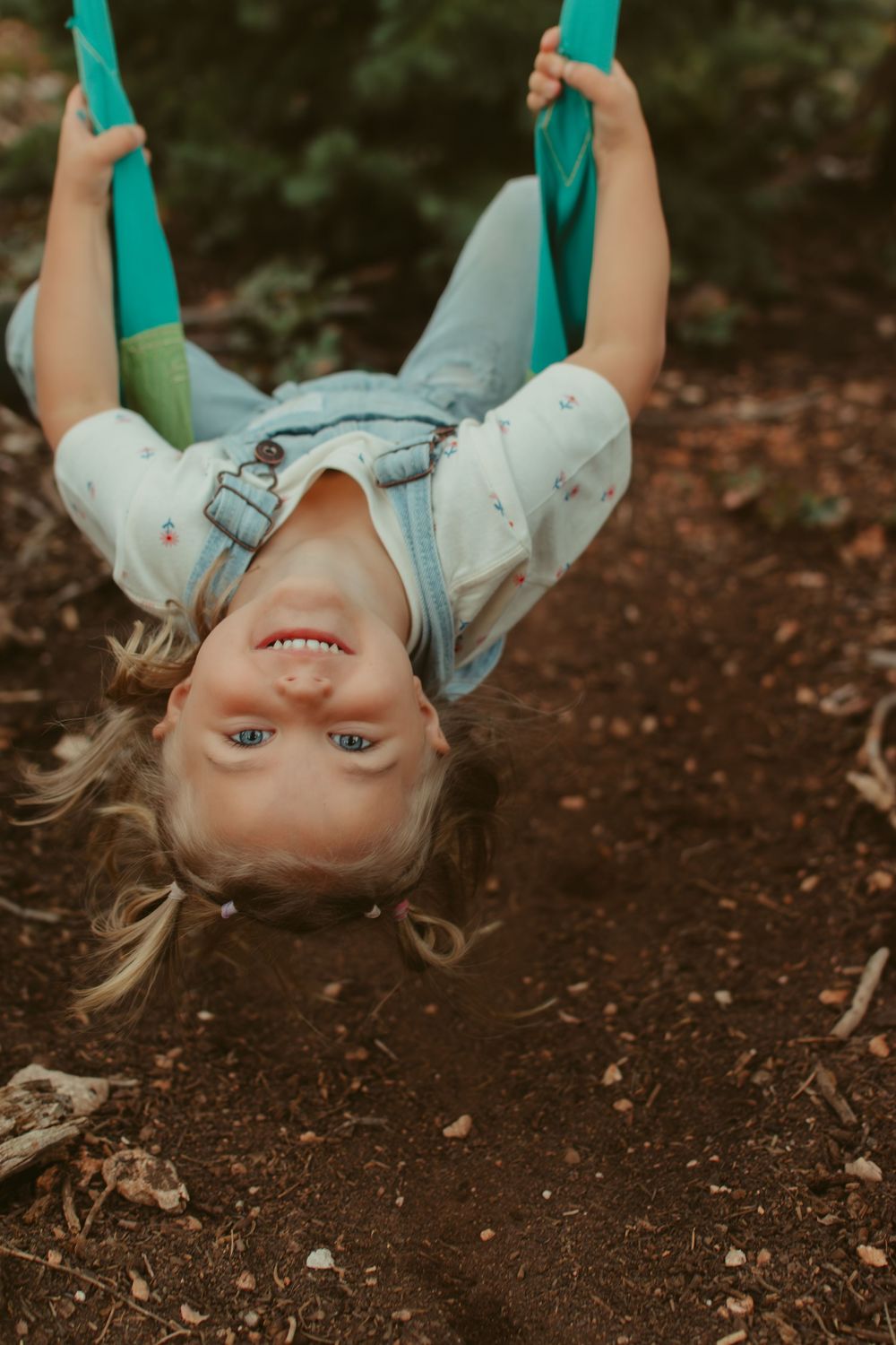 Young girl hanging upside down on a teal swing, smiling joyfully outdoors in a natural woodland setting.