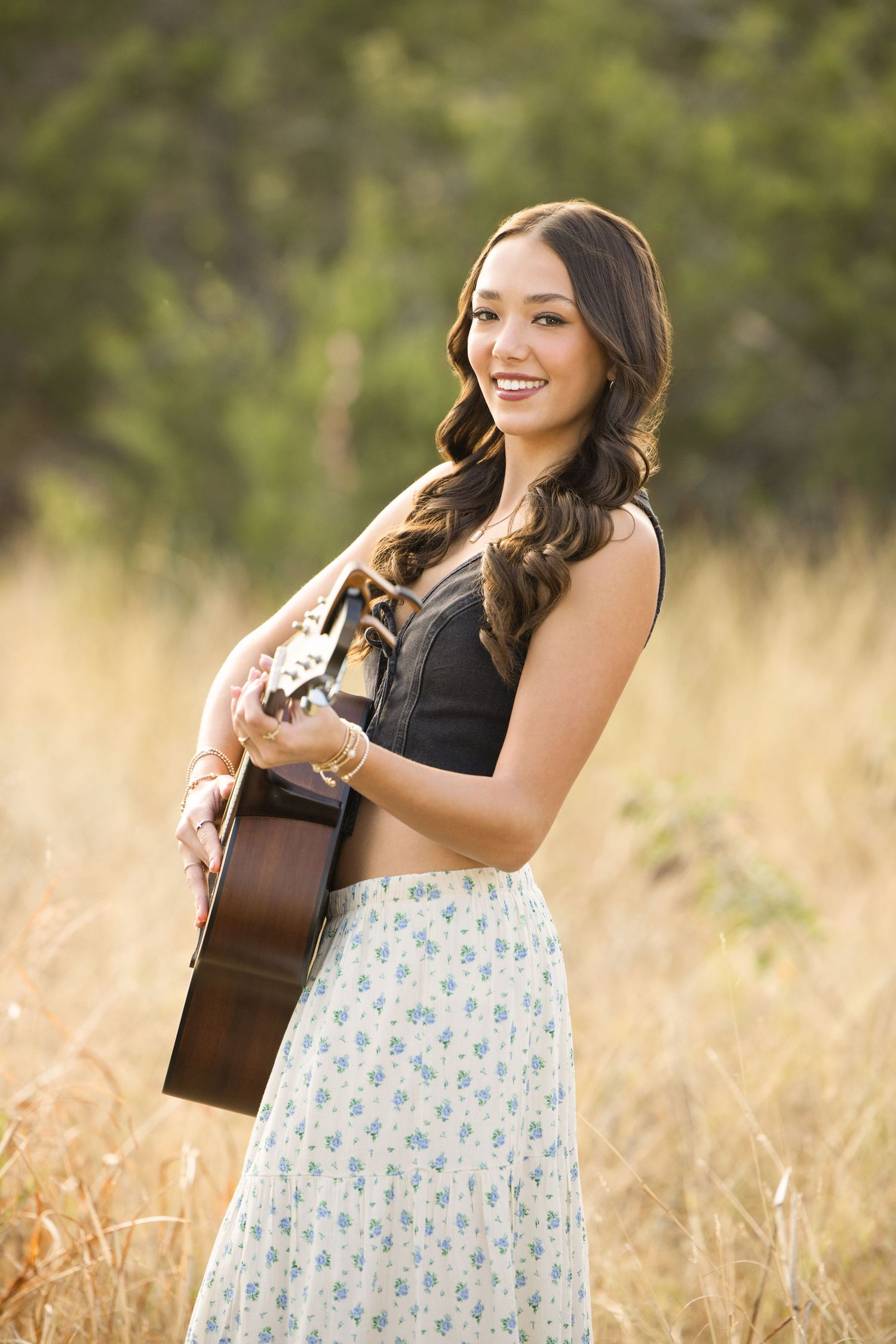 Musician in floral skirt plays acoustic guitar while standing in sunlit golden grass field.