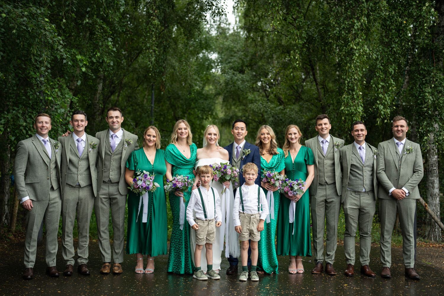 Wedding party poses outdoors with bridesmaids in emerald dresses and groomsmen in gray suits.