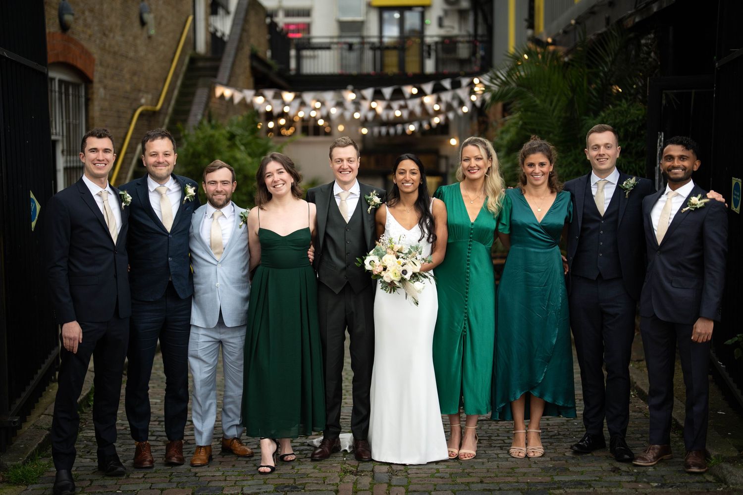 Wedding party poses together with bridesmaids in emerald dresses under string lights in an alley.