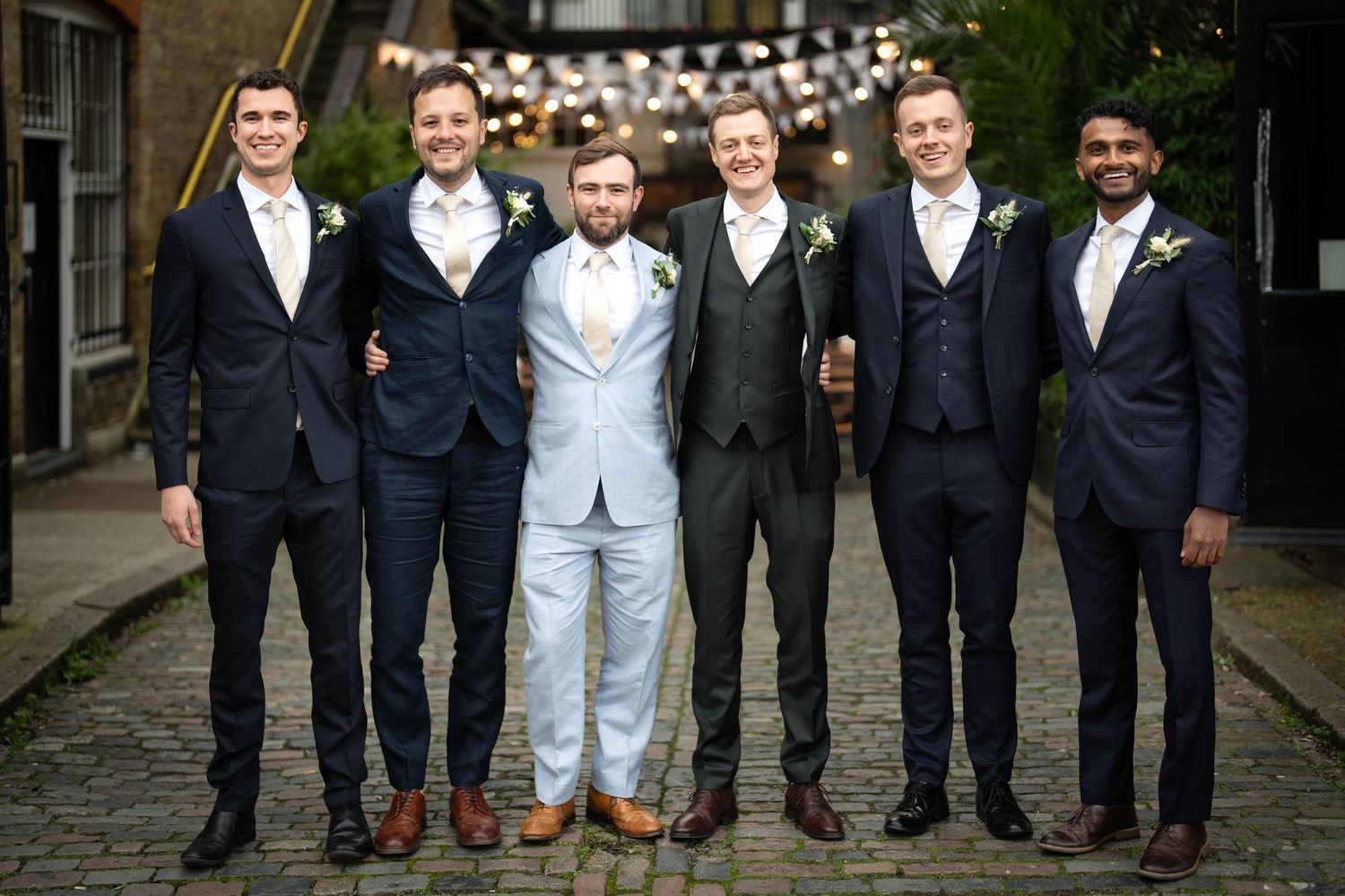 Groomsmen in dark suits pose with one member wearing a light colored suit in a cobblestone alley.