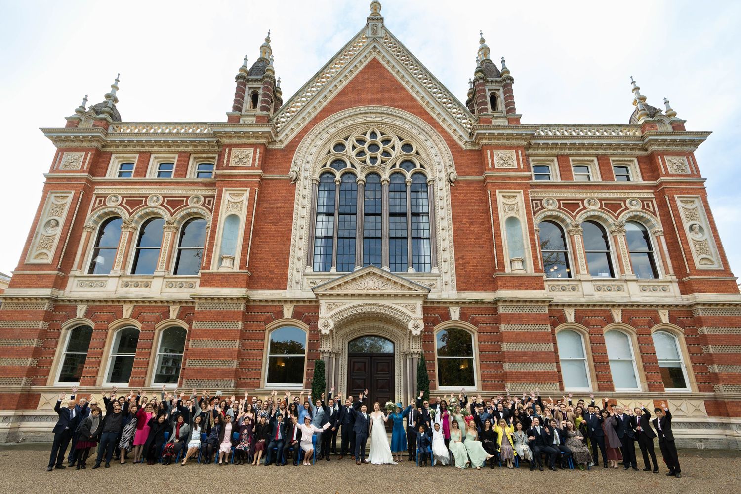 Wedding party poses for group photo outside a grand Victorian red brick building with ornate architecture.