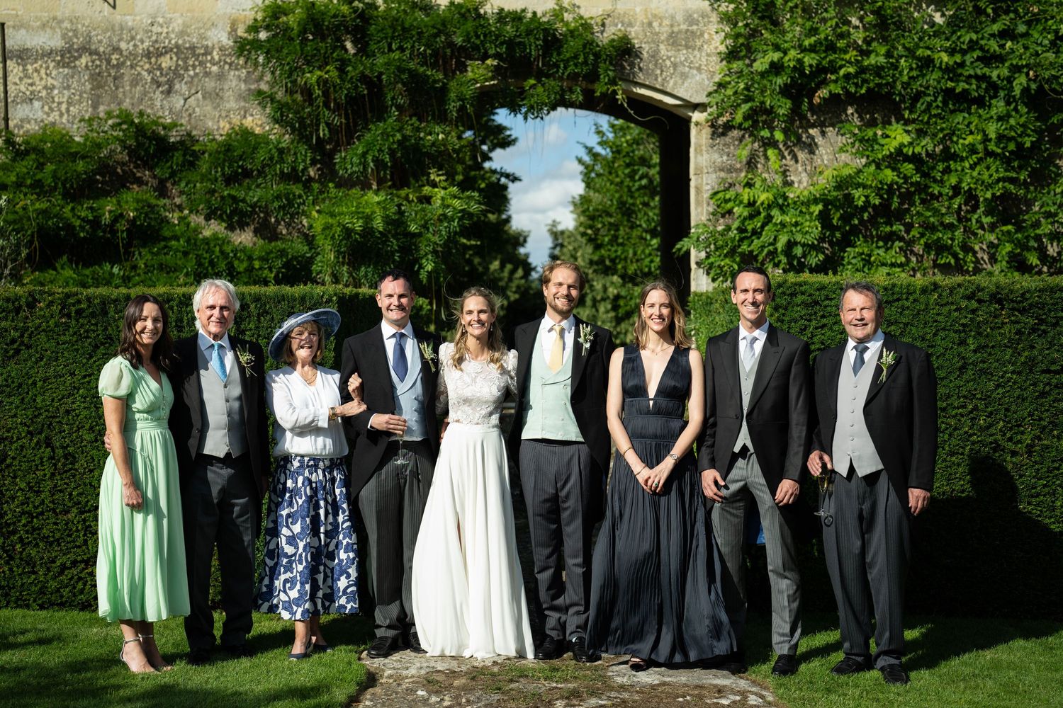 Wedding party poses for formal photo in garden setting framed by a stone archway and hedges.