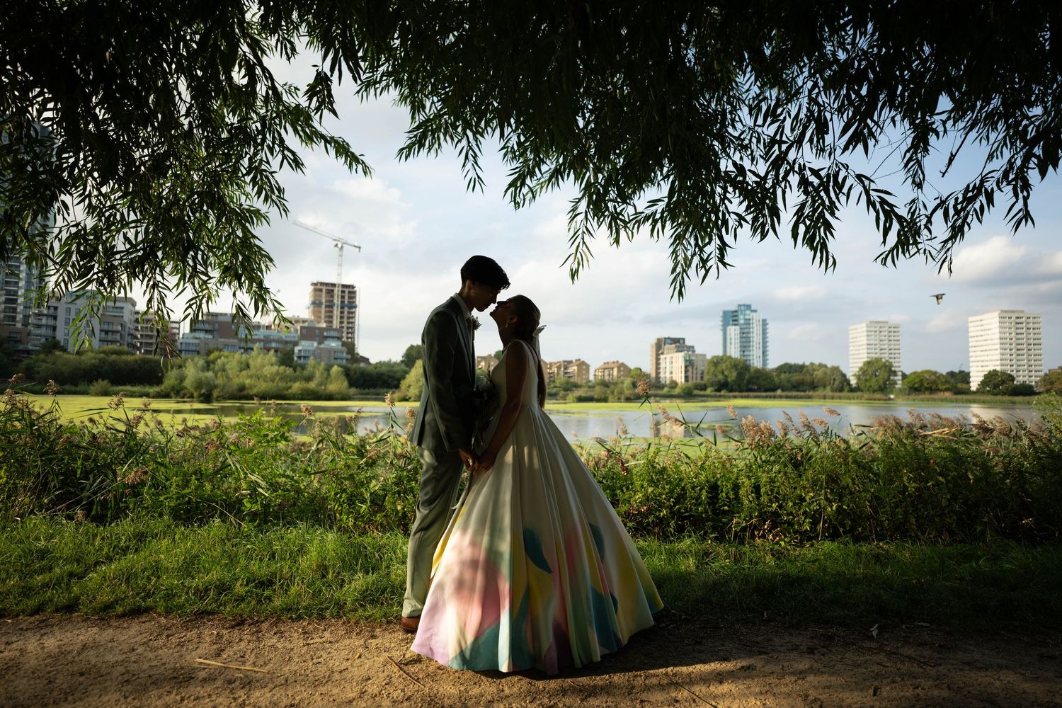 Couple embraces near a lake with city skyline visible in background during golden hour.