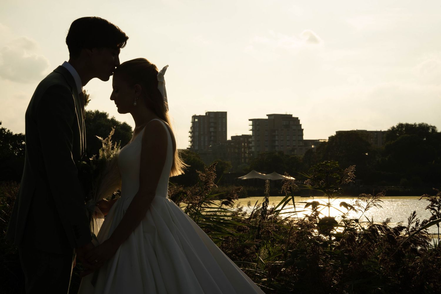 Silhouetted couple share an intimate moment at sunset with cityscape and water in background.