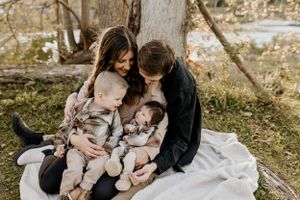 Parents snuggling with twin babies on white blanket in outdoor setting.