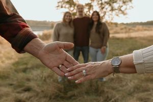 Close-up of hands reaching to hold each other with engagement ring visible, silhouettes standing in golden field at sunset.