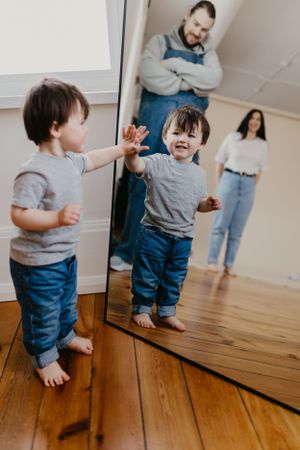 Toddler playing with reflection in full-length mirror while parents watch from background.