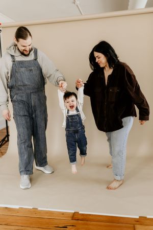 Family in matching denim overalls swinging child between them on wooden floor.