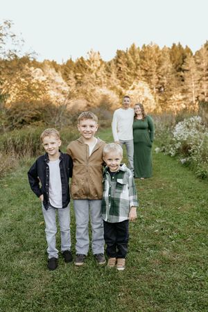 Three children stand together on green grass with autumn trees in the background during a fall photo session.